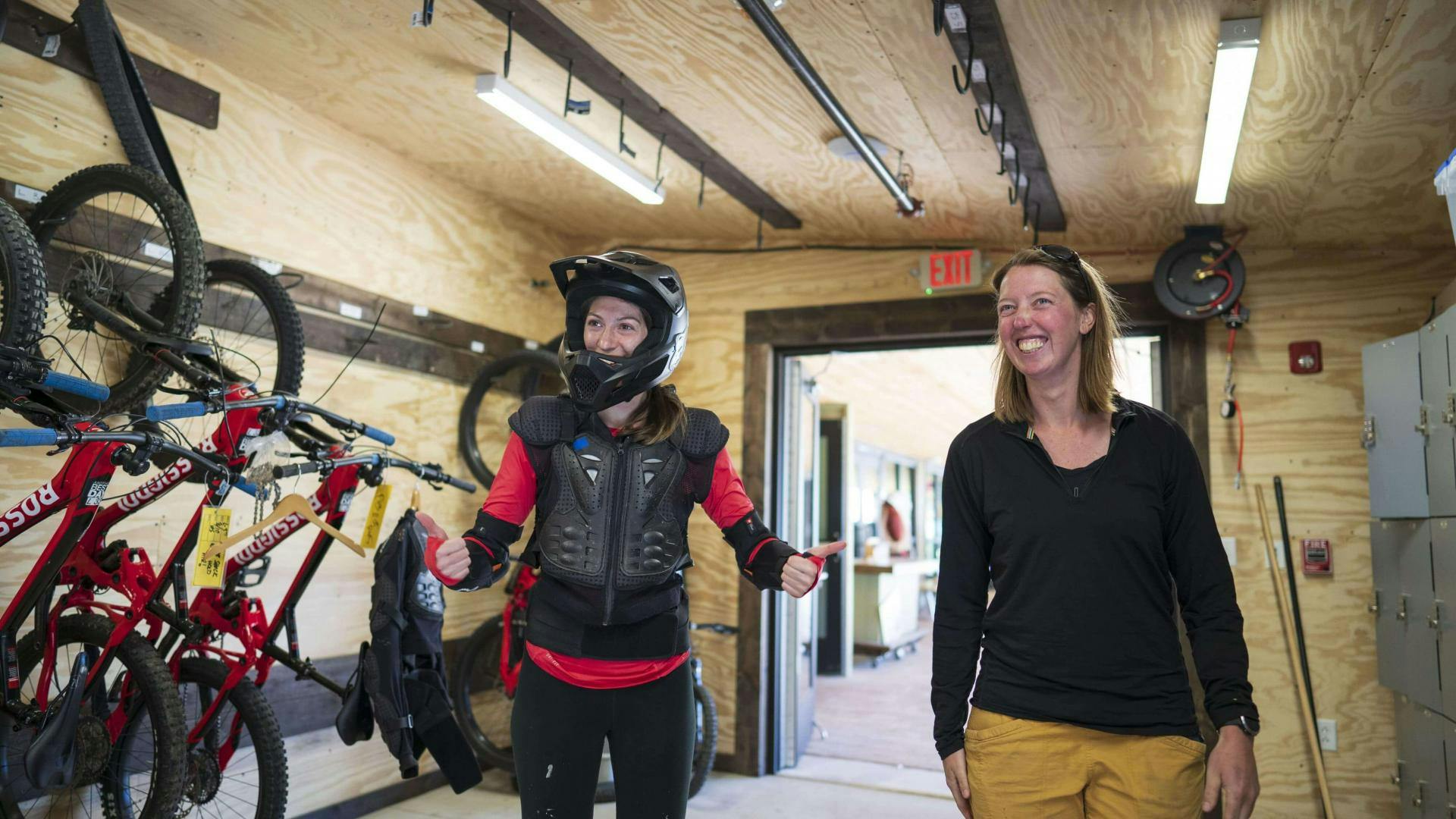 Woman in mountain biking safety gear giving a thumbs up in bike shop next to TAOS employee.