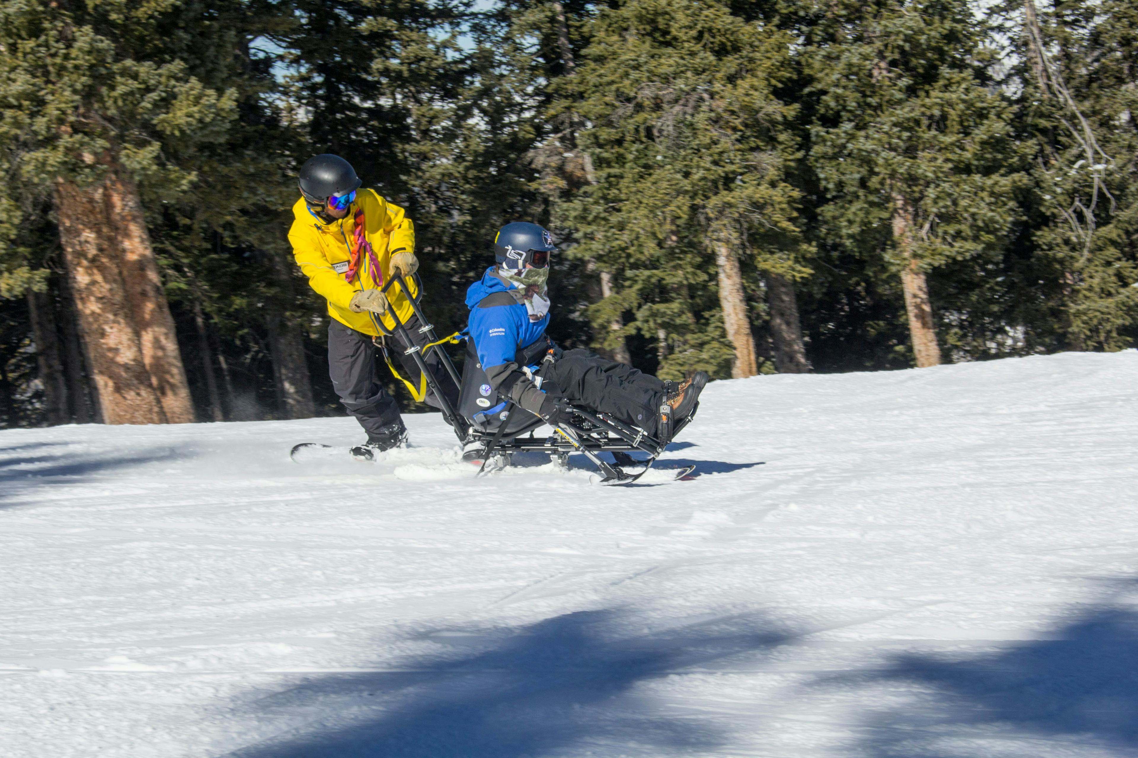 Adaptive skier on a ski chair heads down the mountain with the help of a snow sports instructor